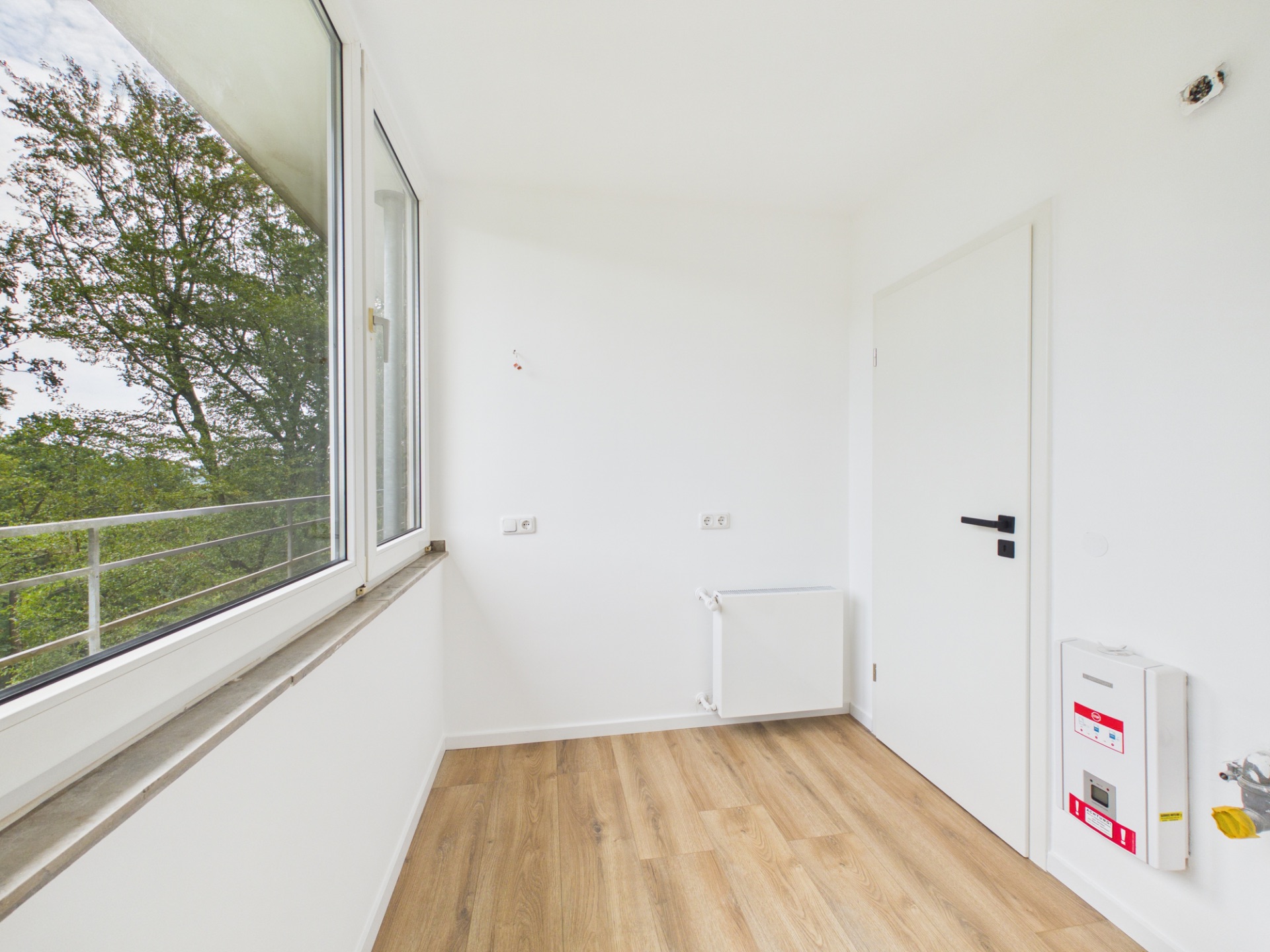 Heller Raum mit großem Fenster, Holzboden, Heizkörper und weißer Tür.