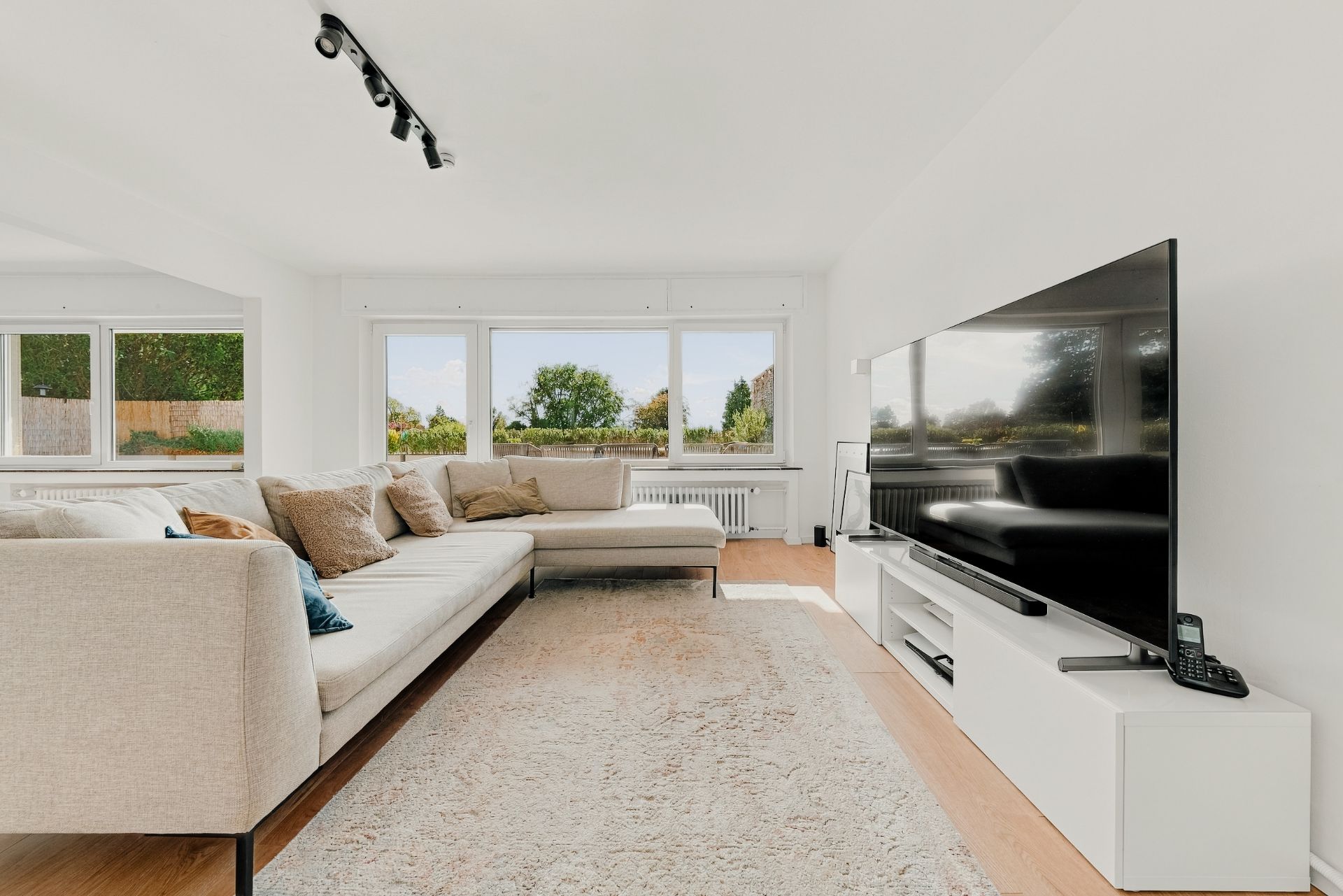 Helles Wohnzimmer mit großem Sofa, Teppich, Fernseher und Fenstern mit Blick ins Grüne.