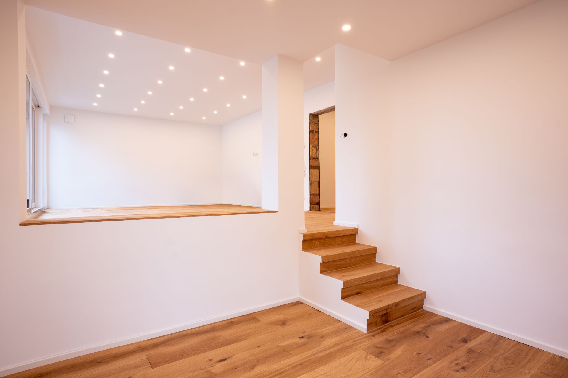 Heller Raum mit Holzboden, kleinen Treppenstufen und vielen Deckenleuchten.
