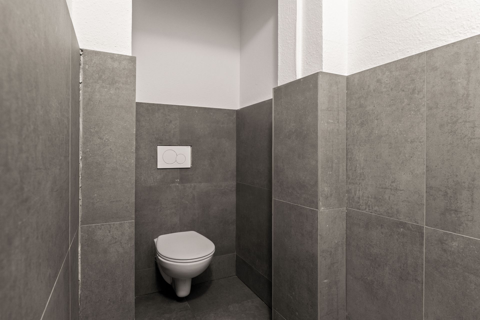 Ein kleines WC mit Wandtoilette und grauen Fliesen an Wänden und Boden.