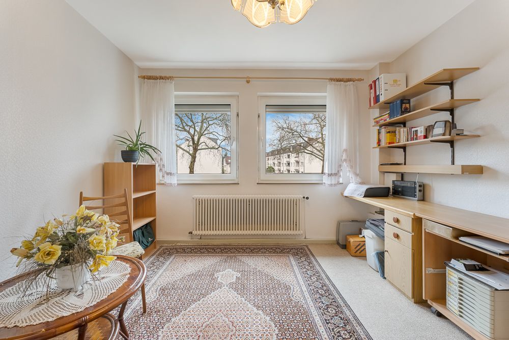 Helles Arbeitszimmer mit Teppich, Schreibtisch, Regal, Tisch, zwei Fenstern und Pflanzen.