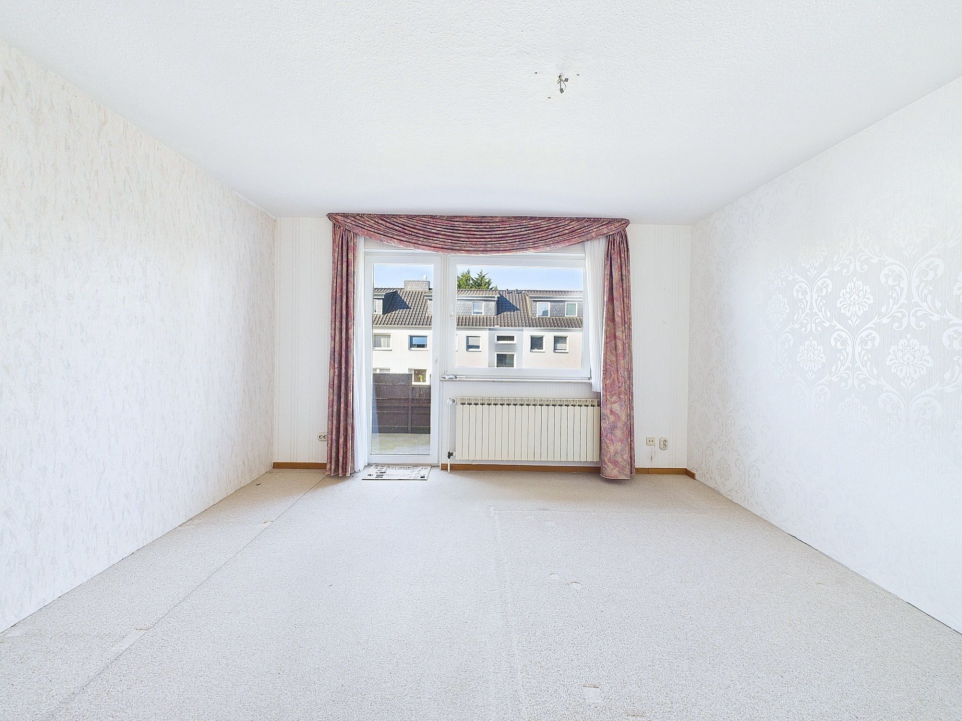 Helles, leeres Zimmer mit Teppichboden, Fenster und Tür zum Balkon, rosa Vorhänge.
