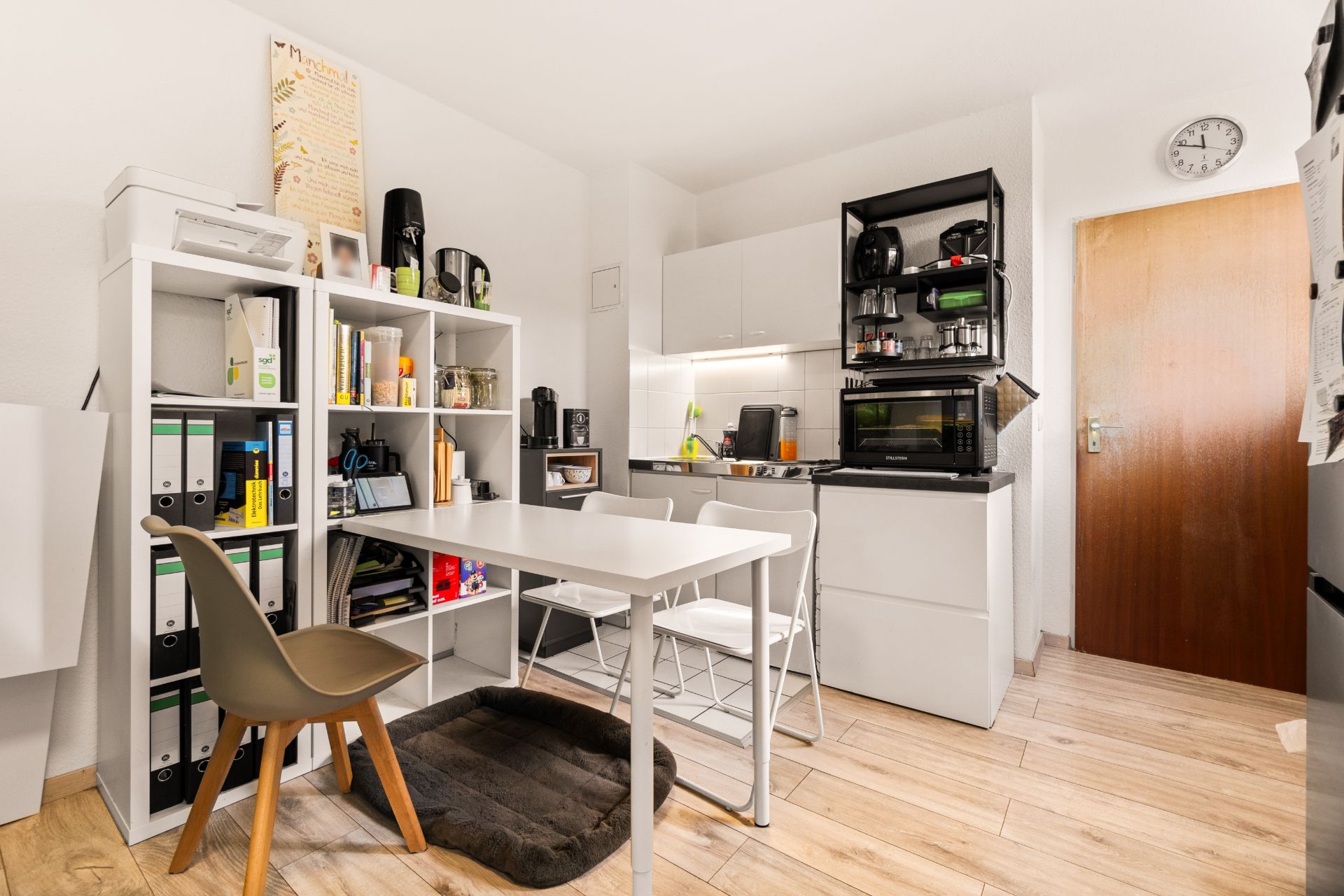 Kleine Küche mit Esstisch, Stühlen, Regal mit Ordnern und Küchengeräten, Holzboden.