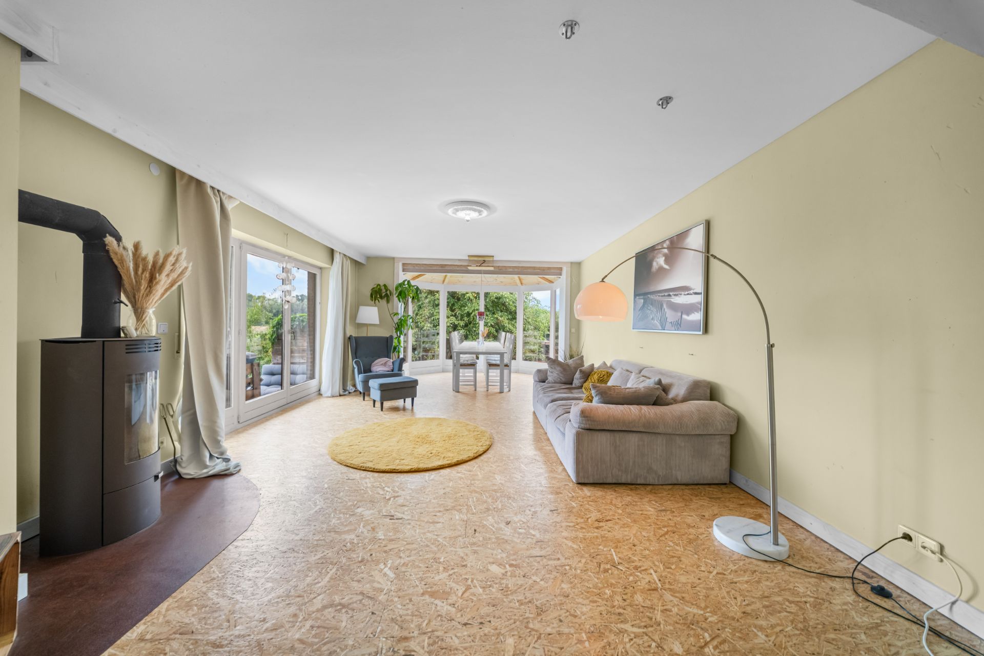 Helles Wohnzimmer mit Sofa, Teppich, Stehlampe, Kaminofen und großem Fenster mit Blick ins Grüne.