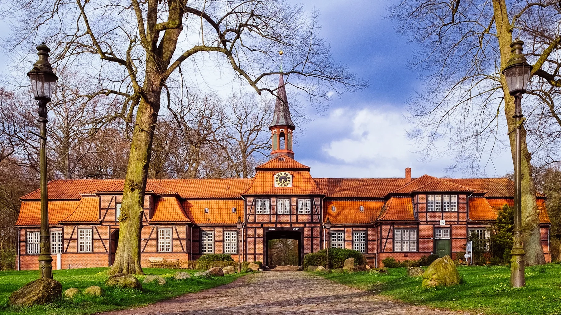 Großes Fachwerkhaus mit rotem Dach und Uhrturm, umgeben von Bäumen und Laternen.