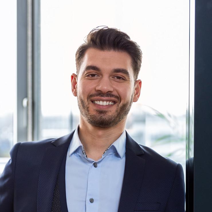 Ein lächelnder Mann im Anzug steht vor einem Fenster in einem Innenraum.