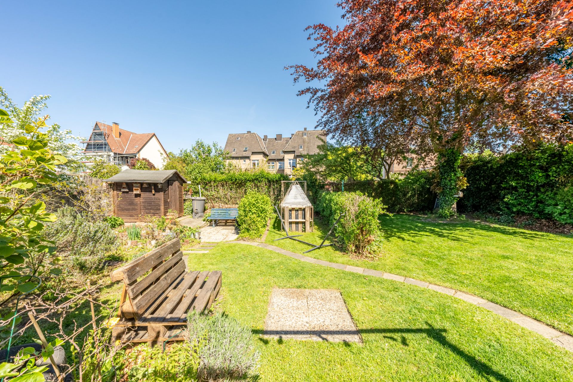 Garten mit Rasen, Bäumen, Holzbank, kleinem Schuppen und Gartenwegen bei sonnigem Wetter.