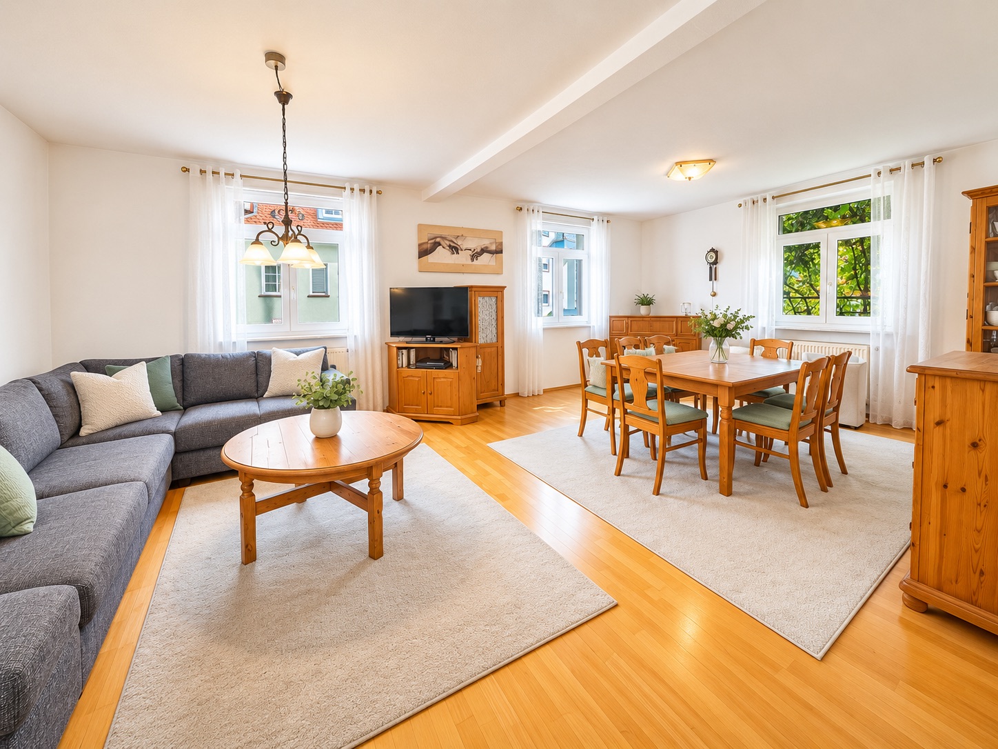 Helles Wohnzimmer mit Ecksofa, Holztisch, Esstisch und großen Fenstern.