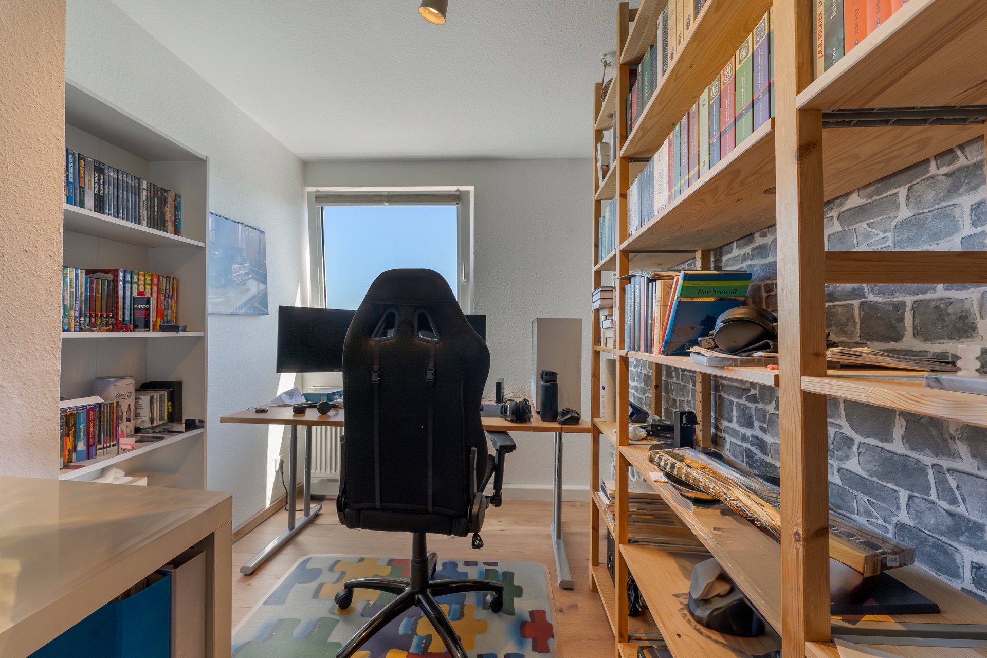 Heimarbeitszimmer mit Schreibtisch, Stuhl, Bücherregalen und Computer vor einem Fenster.