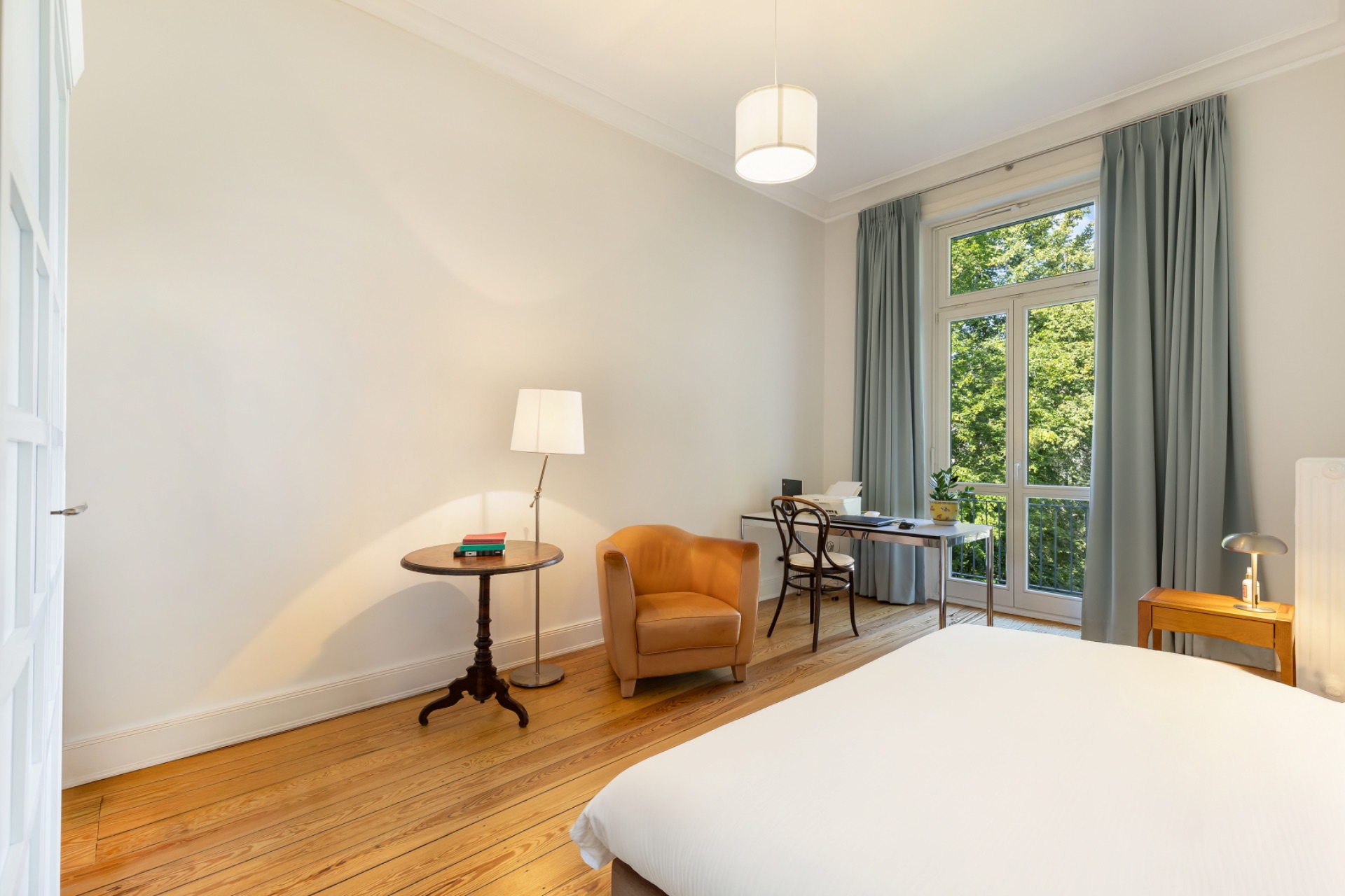 Helles Schlafzimmer mit Sessel, Schreibtisch, großem Fenster und Vorhängen.