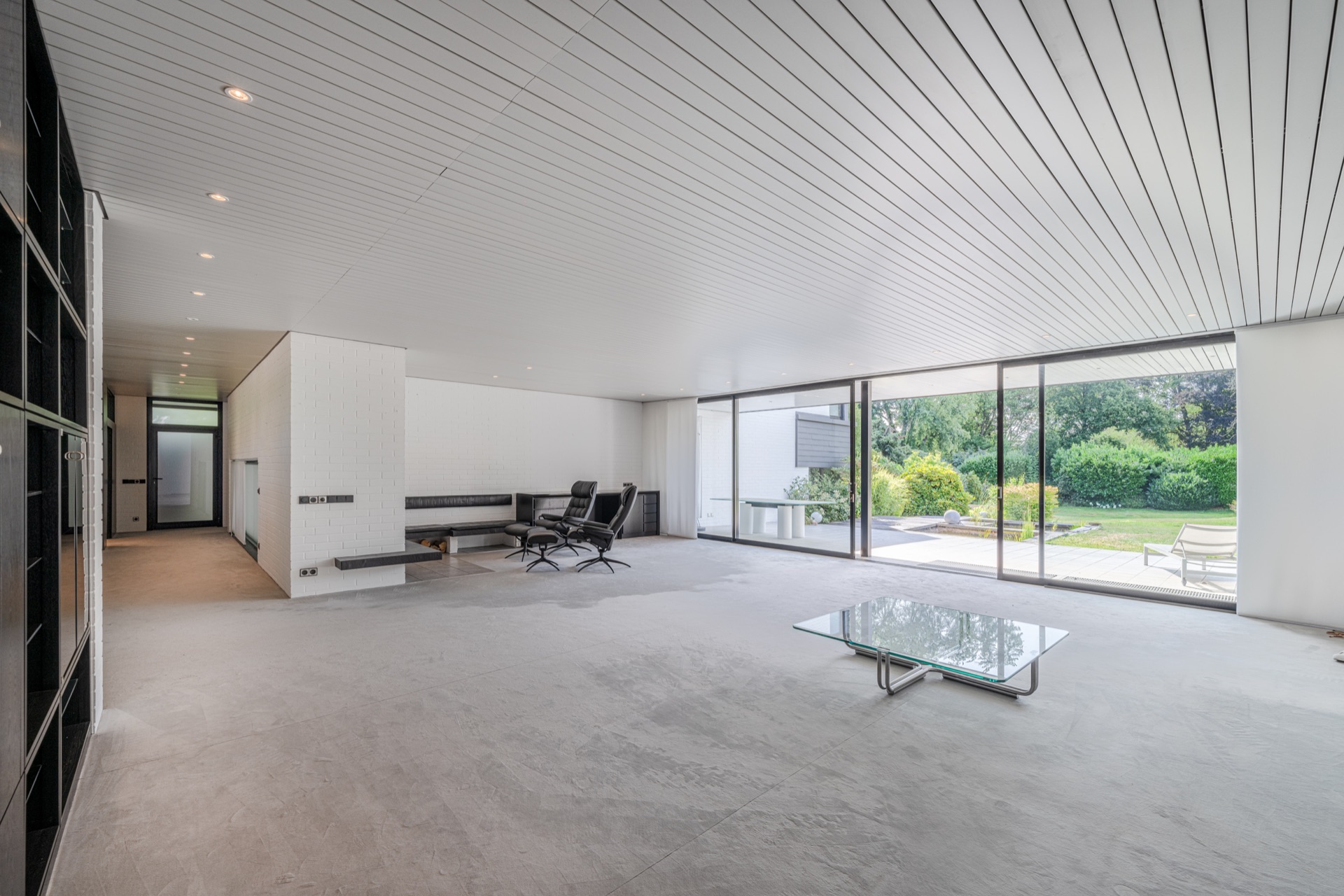 Großes, helles Wohnzimmer mit Glastisch, Sesseln und Blick in den Garten durch große Fenster.