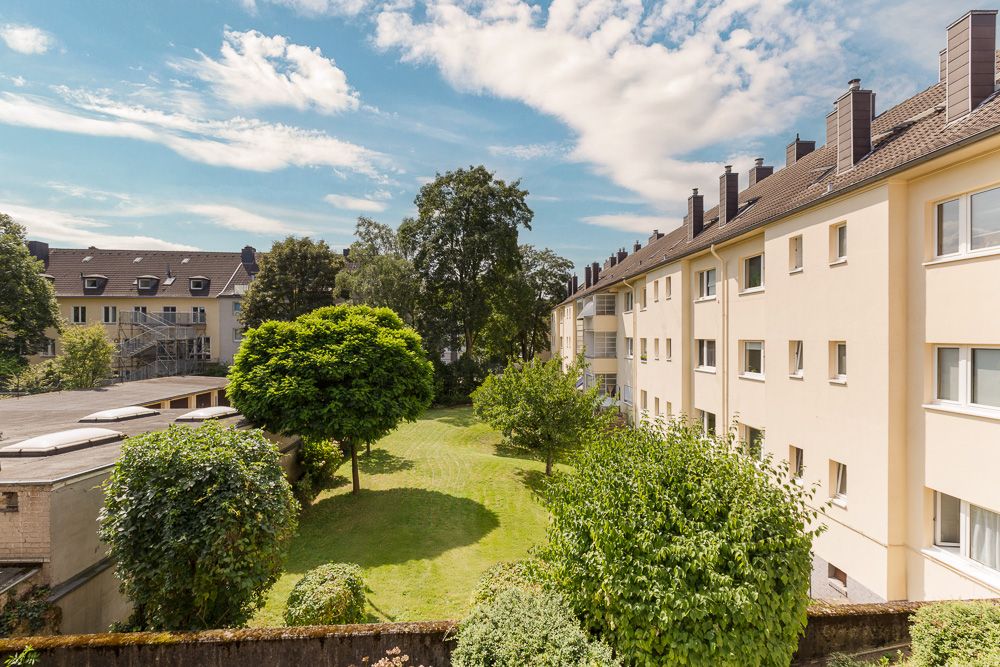 Grüne Wohnanlage mit Rasen, Bäumen und hellen Mehrfamilienhäusern bei blauem Himmel.