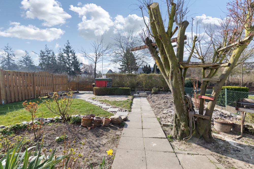 Gepflasterter Weg durch Garten mit Baum, Beeten, Rasen und Holzzaun bei Tageslicht.
