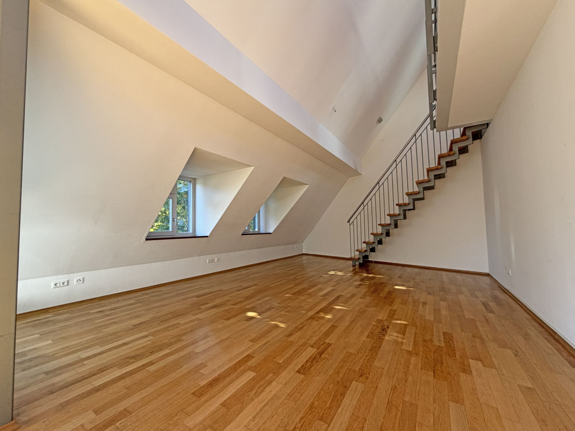 Helles Dachgeschosszimmer mit Holzfußboden, schräge Fenster und Treppe zur oberen Etage.