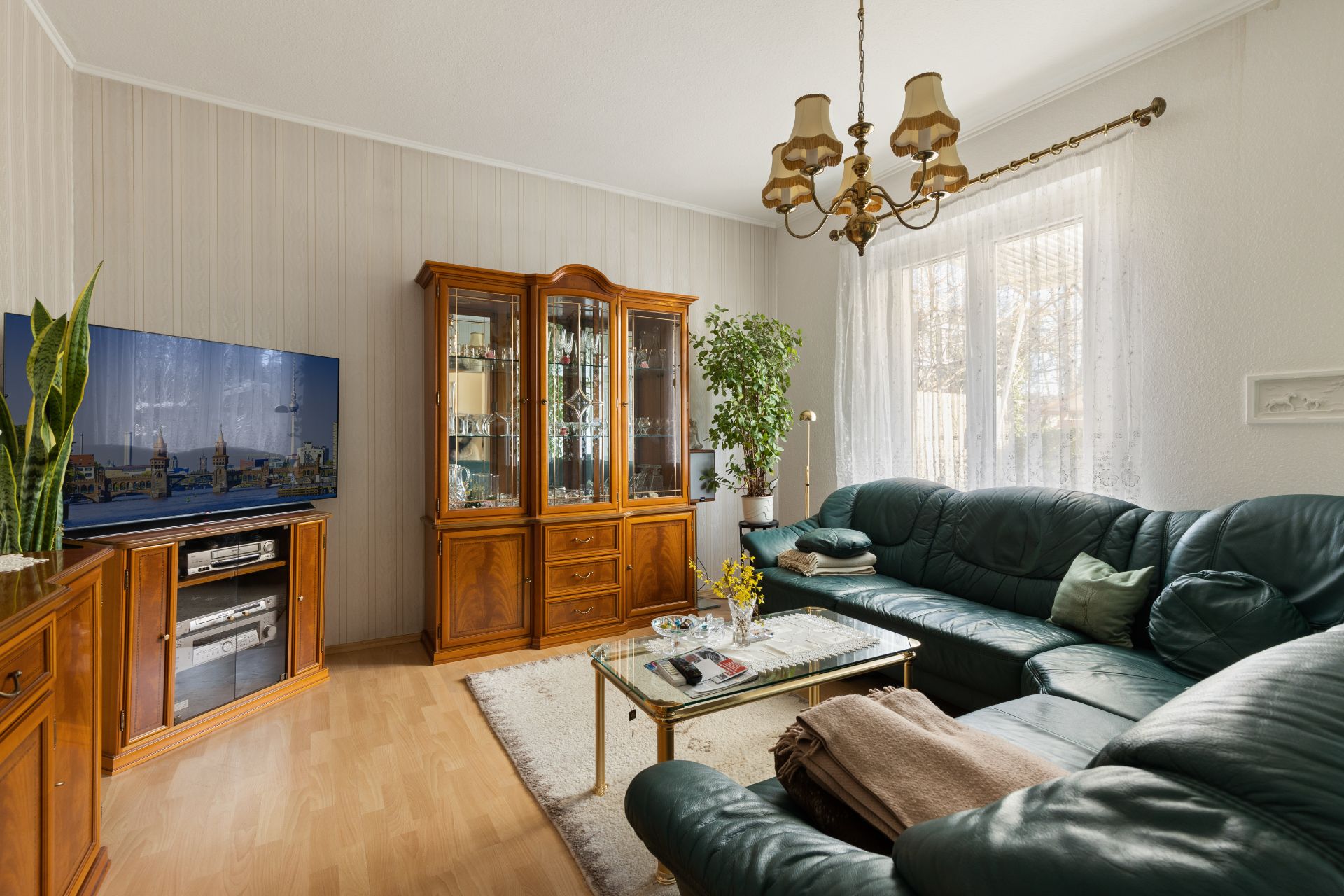 Wohnzimmer mit grüner Ledercouch, Glastisch, Schrankwand, Fernseher und großen Fenstern.
