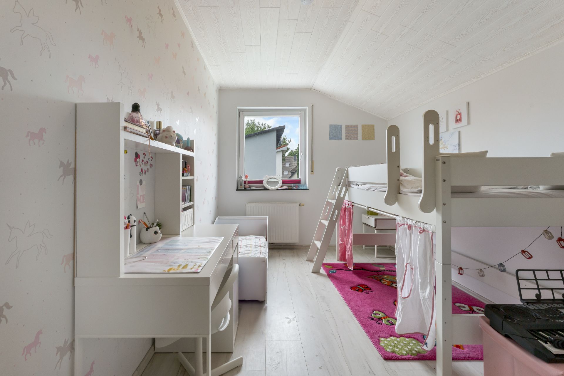 Helles Kinderzimmer mit Hochbett, Schreibtisch und buntem Teppich.
