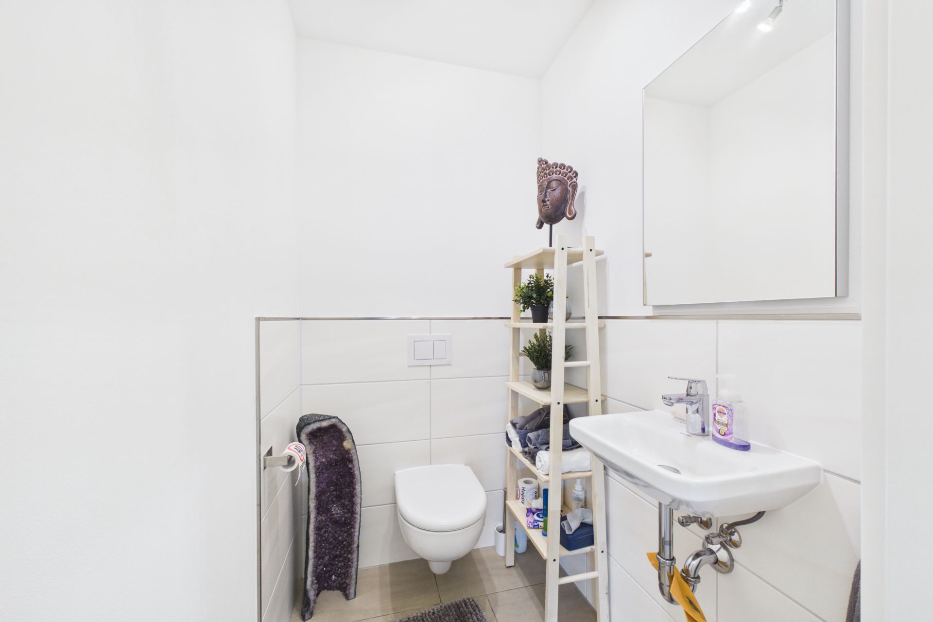 Kleines, helles Badezimmer mit Wandtoilette, Waschbecken, Regal und Spiegel.