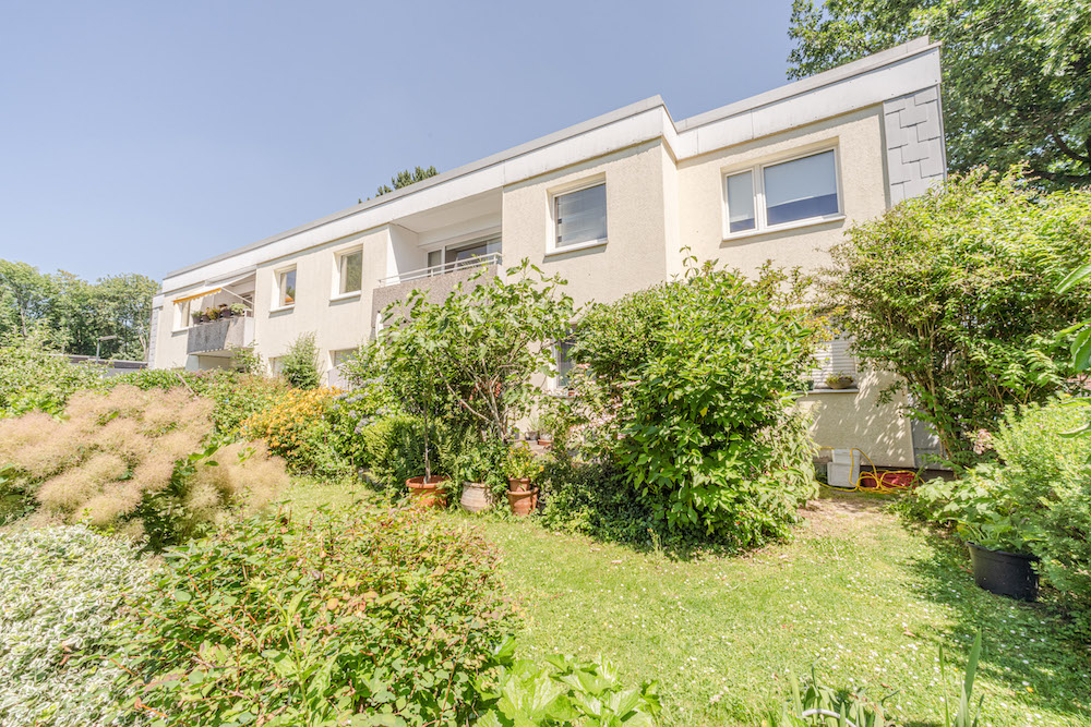 Wohnhaus mit zwei Etagen und Balkon, umgeben von grünem Garten mit Sträuchern und Pflanzen.