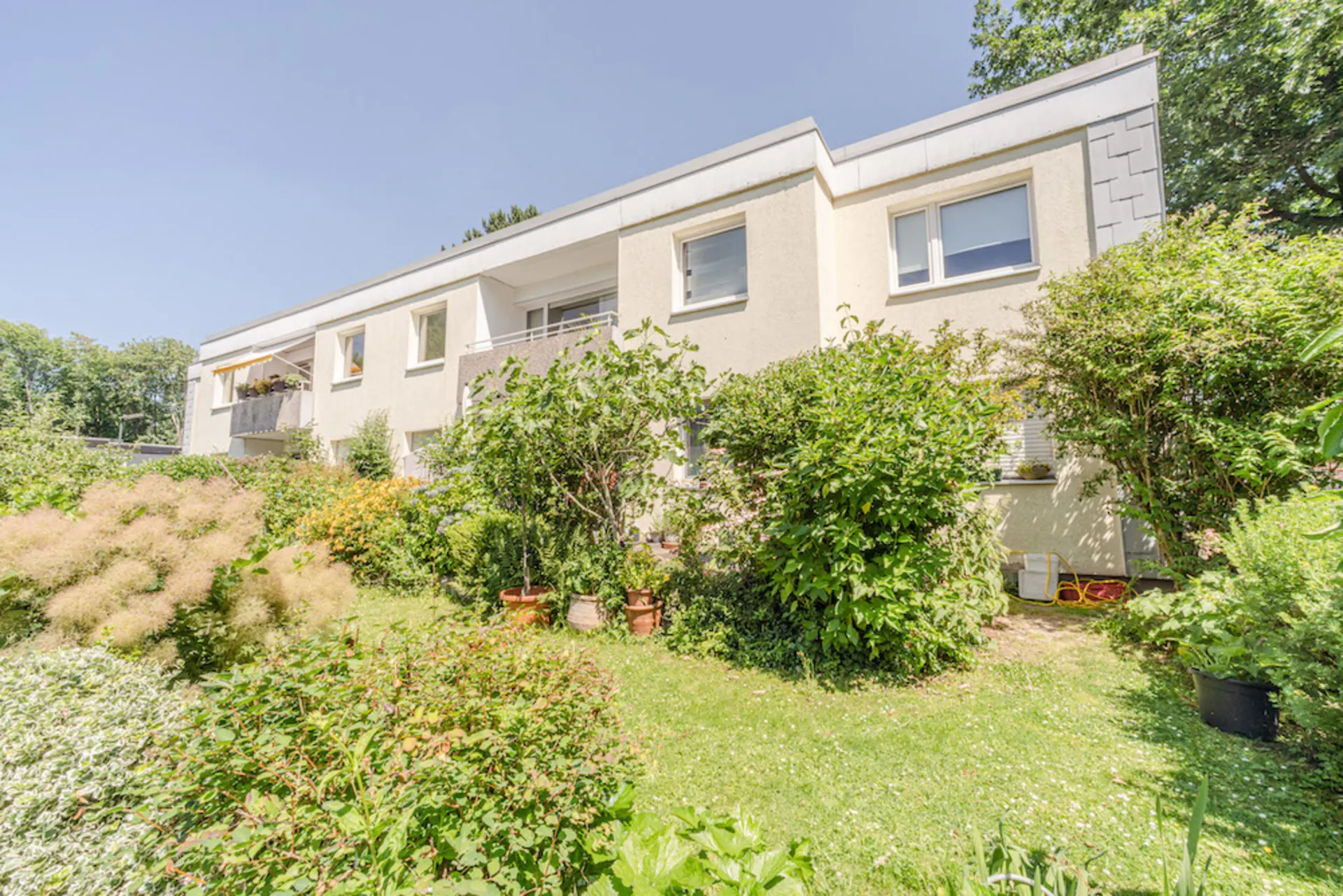 Wohnhaus mit zwei Etagen und Balkon, umgeben von grünem Garten mit Sträuchern und Pflanzen.