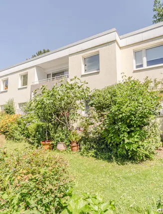 Wohnhaus mit zwei Etagen und Balkon, umgeben von grünem Garten mit Sträuchern und Pflanzen.