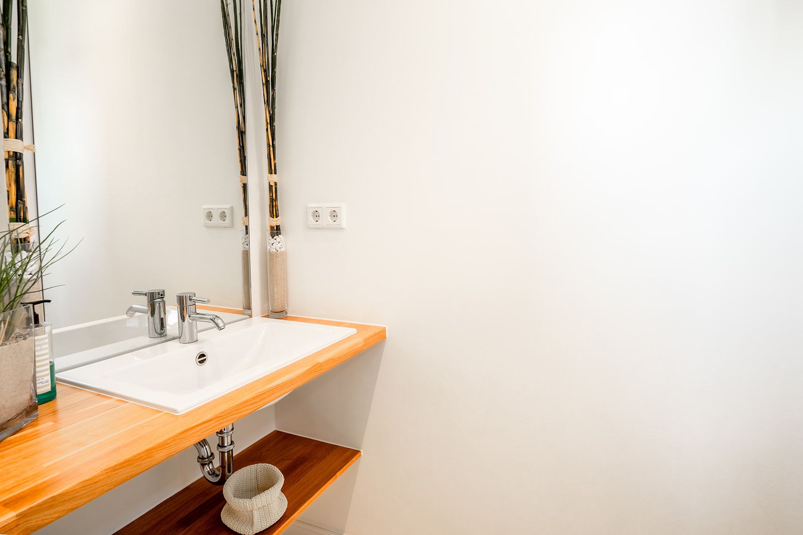 Waschbecken mit Holzablage, Spiegel und Dekozweigen an weißer Wand.
