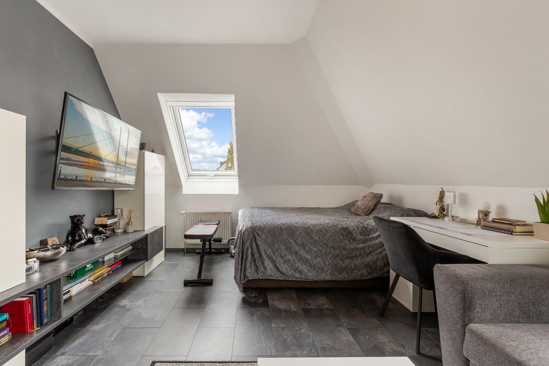 Helles Schlafzimmer mit Dachfenster, Bett, Schreibtisch und Fernseher an der Wand.