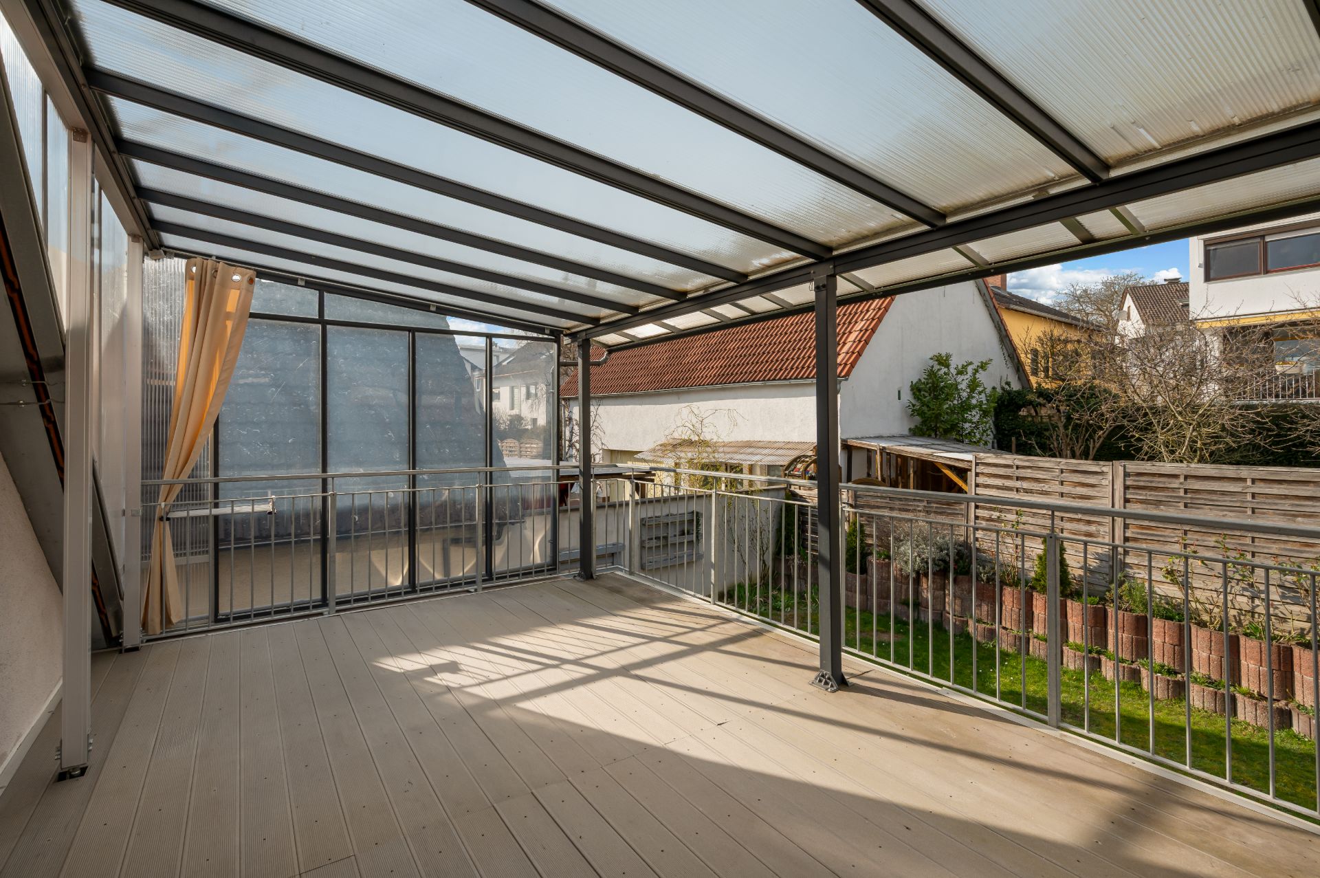 Überdachte Terrasse mit Geländer und Blick auf einen kleinen Garten und Nachbarhäuser.
