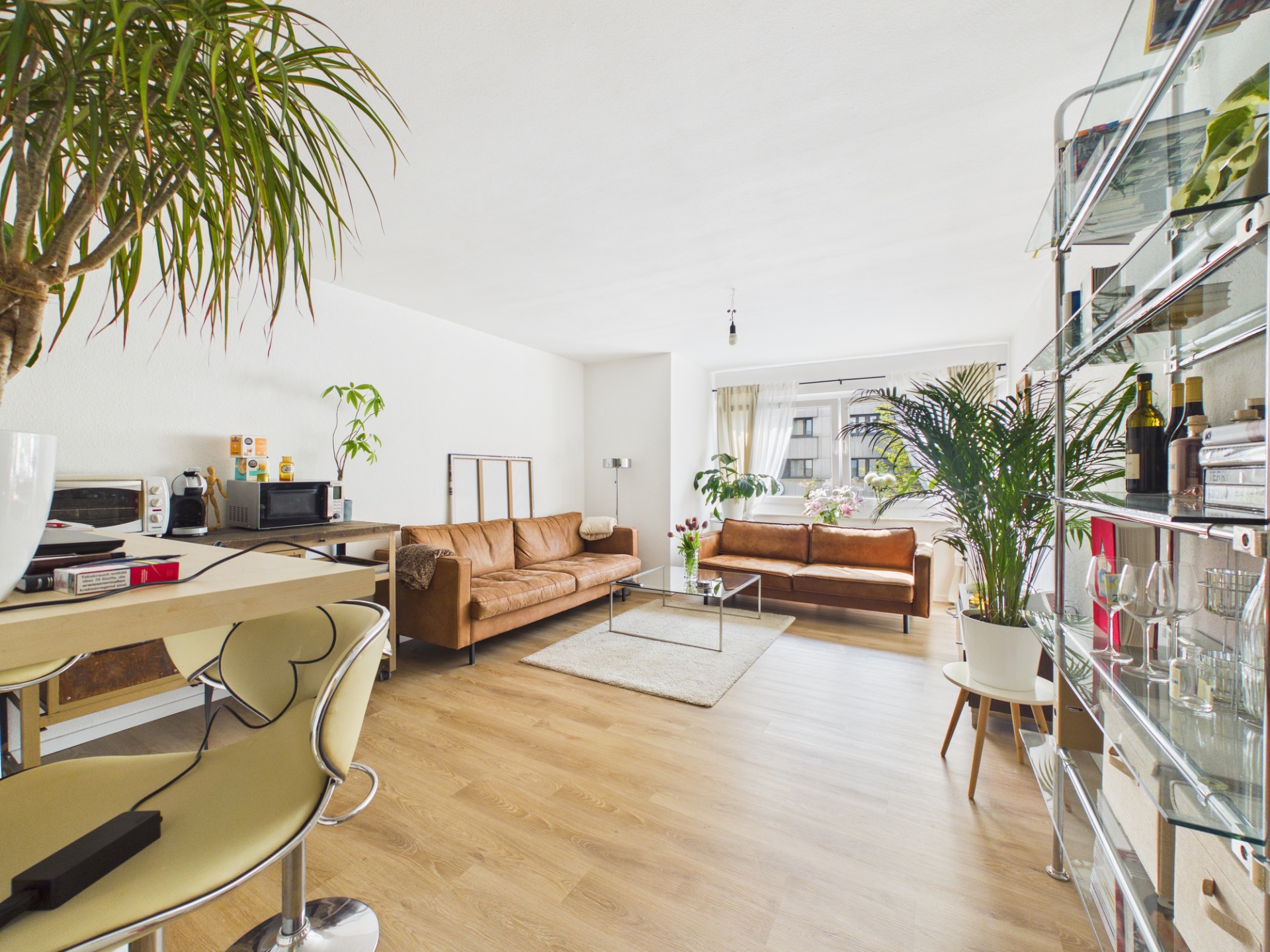Helles Wohnzimmer mit Holzboden, zwei braunen Sofas, Schreibtisch, Pflanzen und Regalwand.