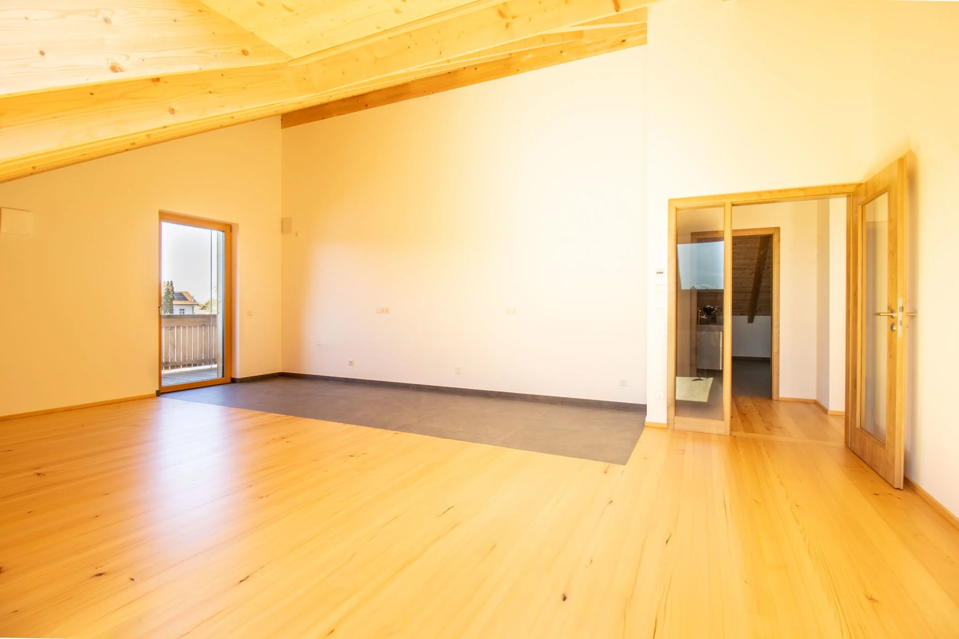Helles, leeres Zimmer mit Holzfußboden, Holzbalkendecke, Tür zum Balkon und Glastür zum Flur.