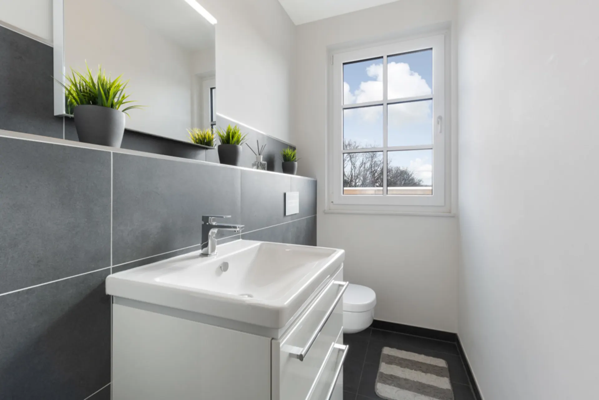 Modernes Badezimmer mit Waschbecken, Toilette, Fenster und drei grünen Pflanzen auf einer Ablage.