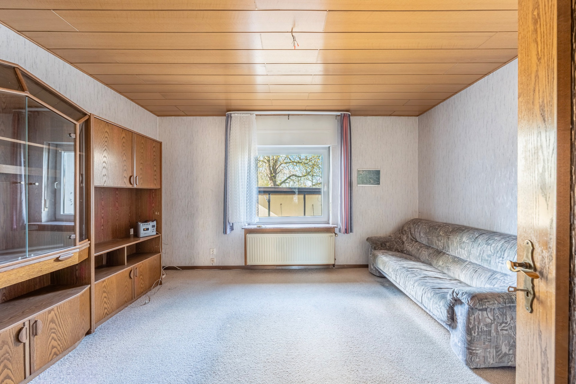 Wohnzimmer mit Holzdecke, Sofa, Schrankwand und Fenster mit Vorhang.