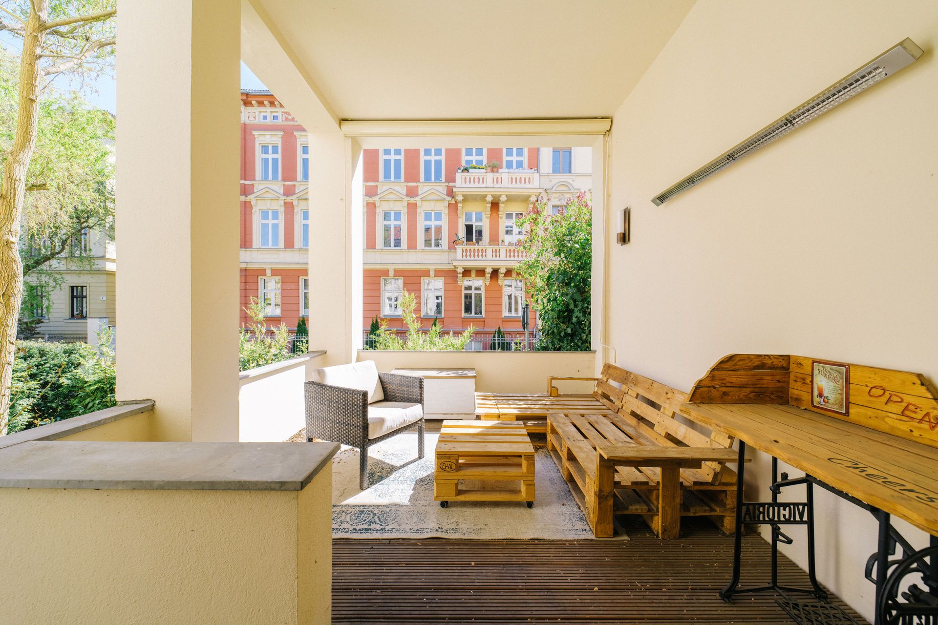Überdachte Terrasse mit Holzbänken, Tisch und Blick auf ein buntes Wohnhaus.