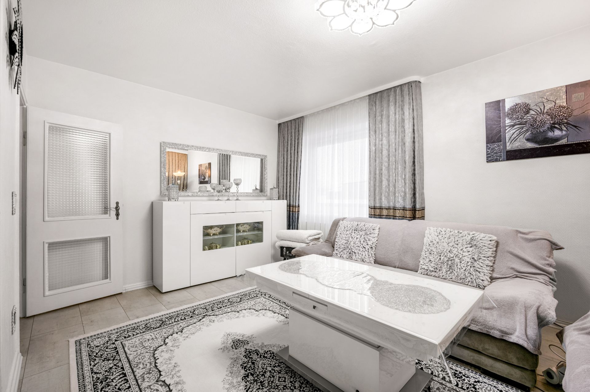 Helles Wohnzimmer mit Sofa, Couchtisch, Teppich, Kommode, Spiegel und großen Fenstern mit Vorhängen.