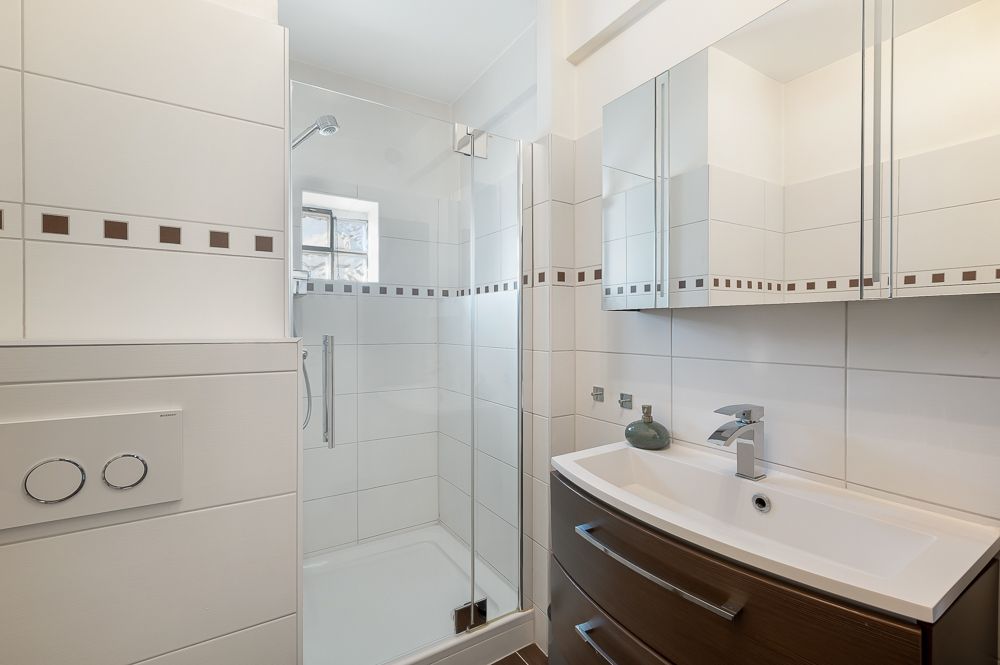 Modernes Bad mit Dusche, Waschbecken und Spiegelschrank, weiße Fliesen mit Dekorstreifen.