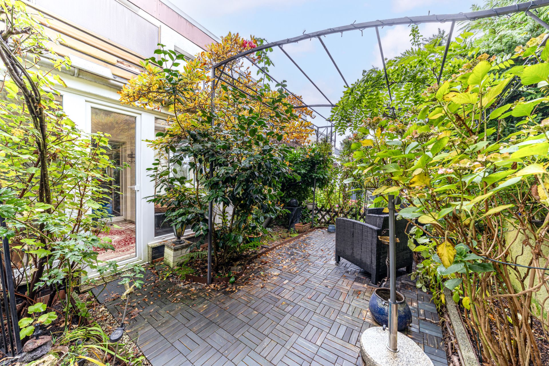 Kleiner Garten mit Terrasse, Pflanzen, Sitzbereich und offenem Rankbogen.