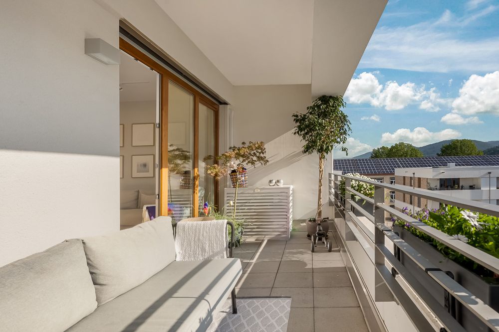 Moderner Balkon mit Sofa, Pflanzen und Blick auf Häuser und Berge bei blauem Himmel.
