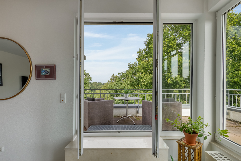 Offenes Fenster mit Blick auf eine Terrasse mit Sitzgruppe und grünem Ausblick.