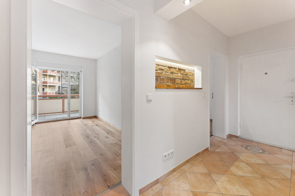 Heller Flur mit Fliesenboden, Sicht auf ein Zimmer mit Holzboden und Balkon, beleuchtetes Ziegelwanddetail.