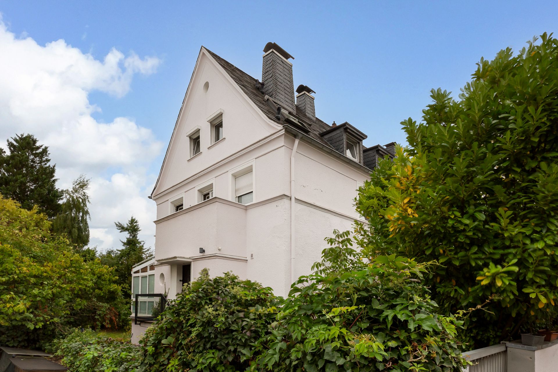 Weißes Wohnhaus mit Satteldach, mehreren Schornsteinen und umgeben von viel Grün.