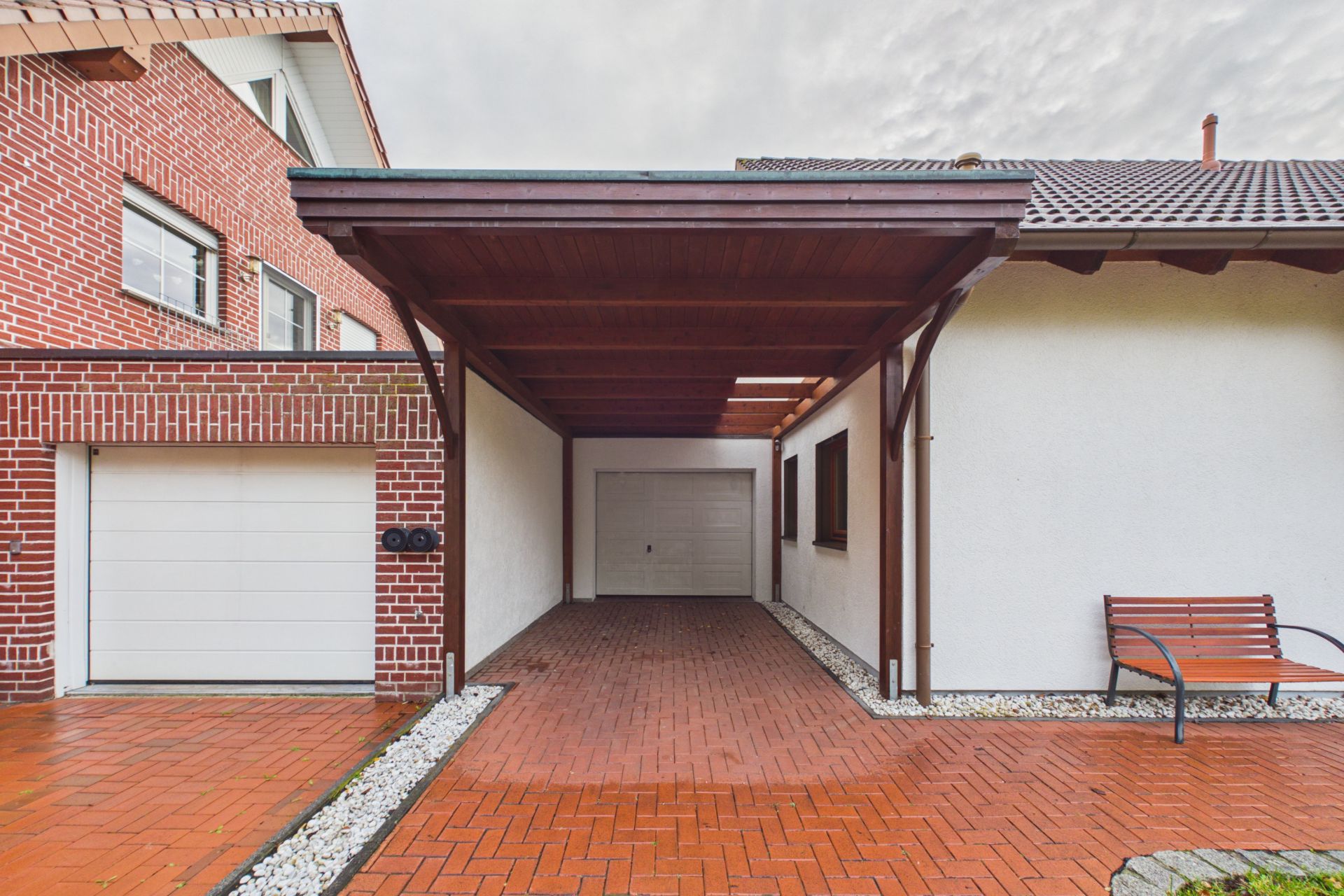 Überdachter Carport mit angrenzender Garage, Backsteinboden und Bank an einem Wohnhaus.