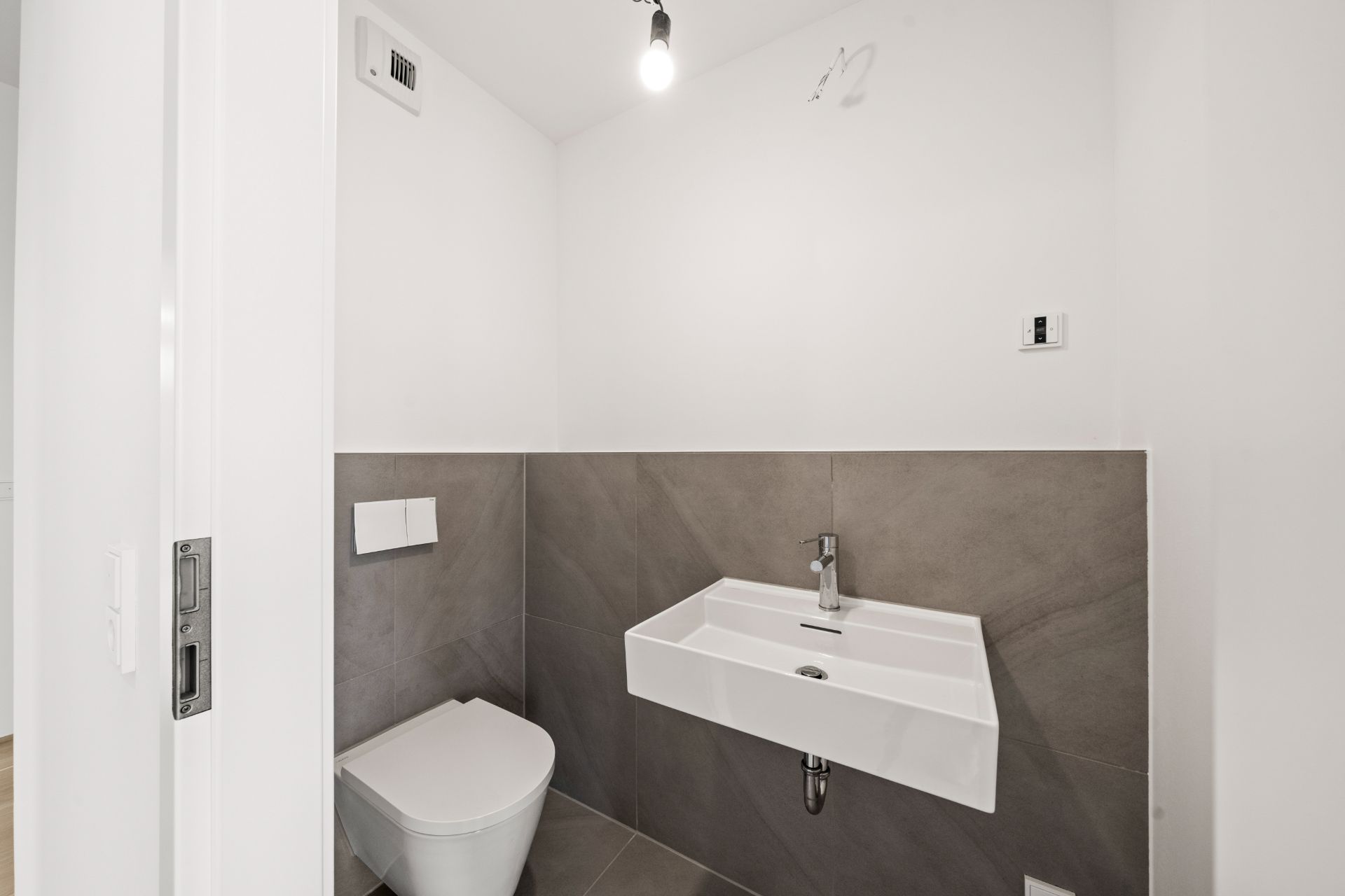 Kleines Bad mit Wandtoilette, rechteckigem Waschbecken und grauen Fliesen.
