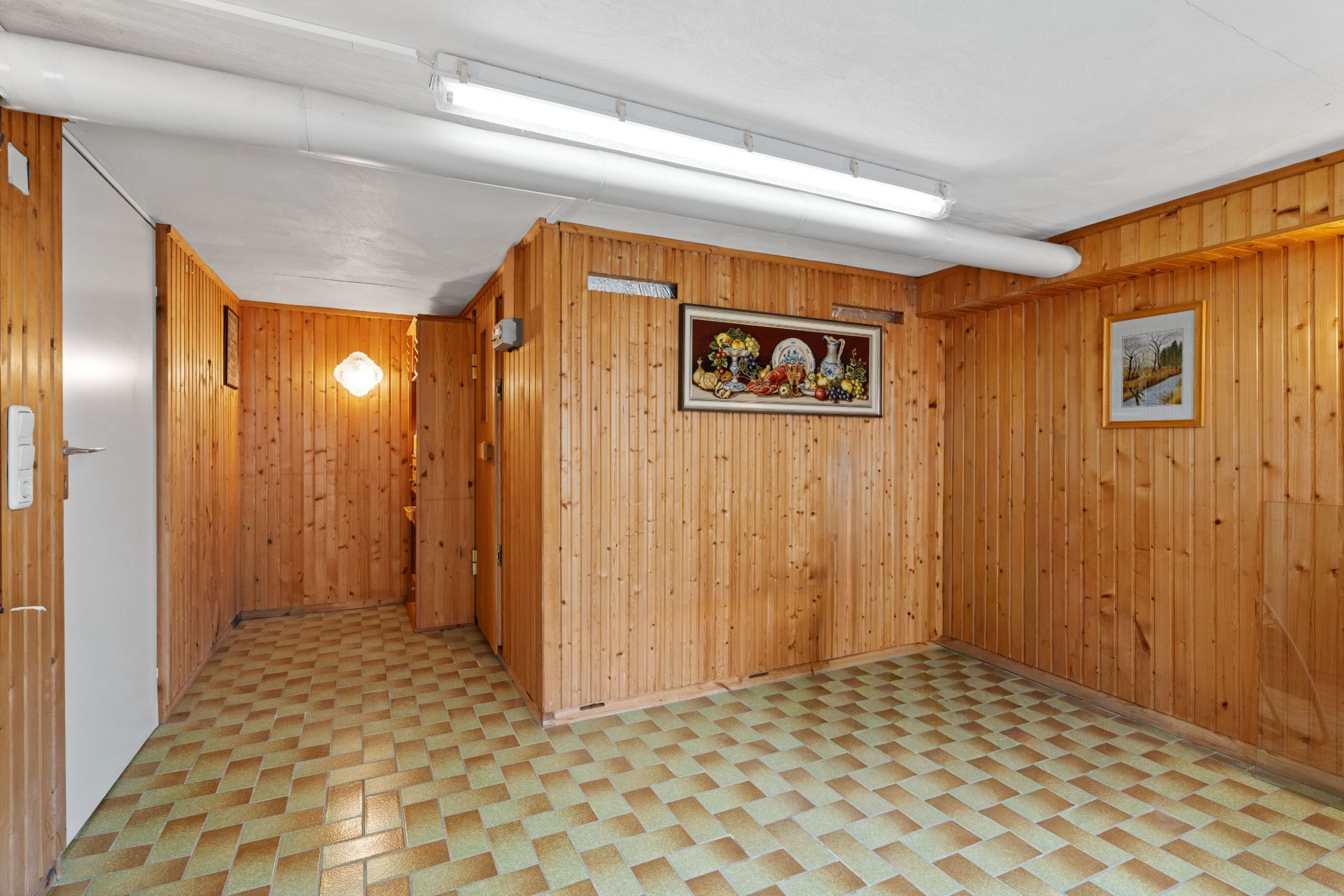 Raum mit Holzvertäfelung, gemustertem Fliesenboden, Deckenleuchte und Wandbildern.