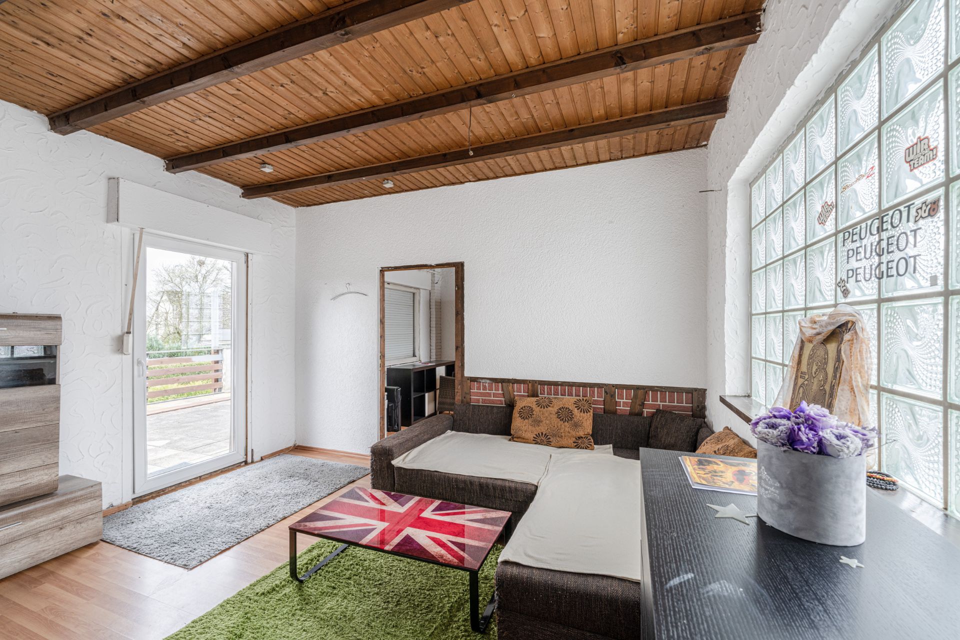 Wohnzimmer mit Ecksofa, Glastisch, Teppich, Holzbalkendecke und großen Glasbausteinfenstern.