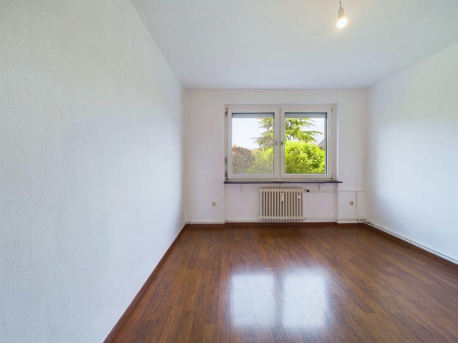 Leerer Raum mit weiß gestrichenen Wänden, Holzfußboden und Fenster mit Blick ins Grüne.