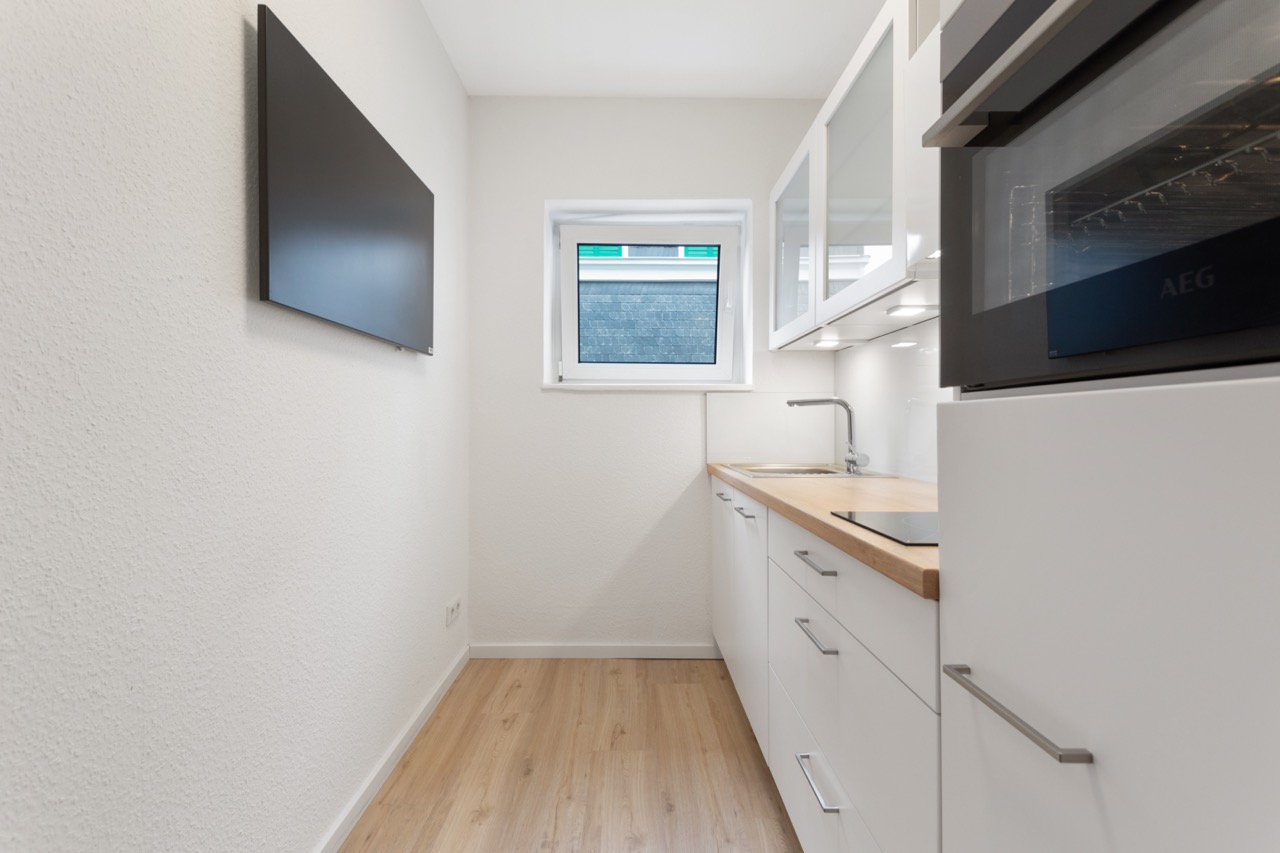 Schmale, moderne Küche mit weißen Schränken, Holzarbeitsplatte, Fenster und Wandbildschirm.