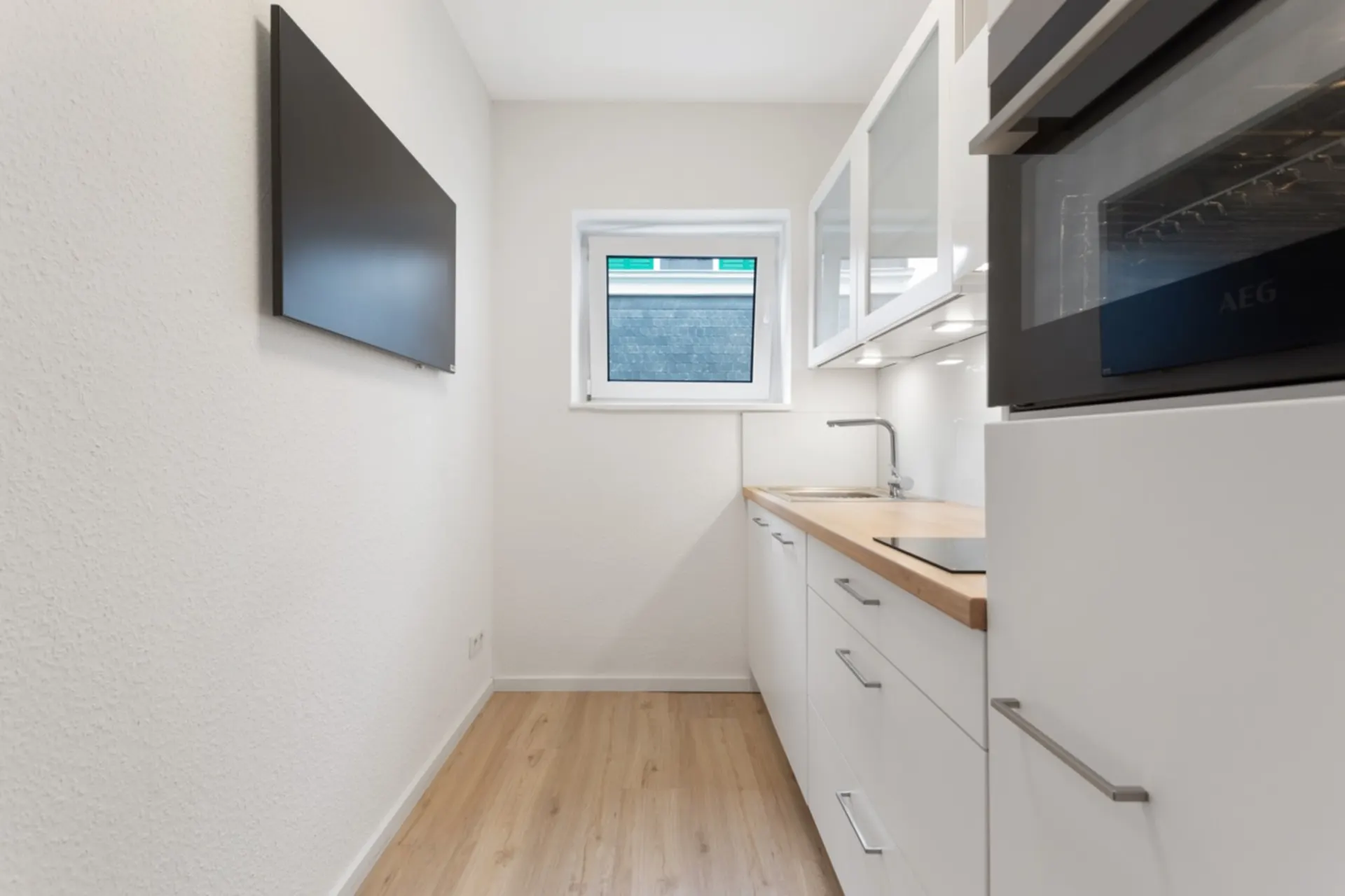 Schmale, moderne Küche mit weißen Schränken, Holzarbeitsplatte, Fenster und Wandbildschirm.