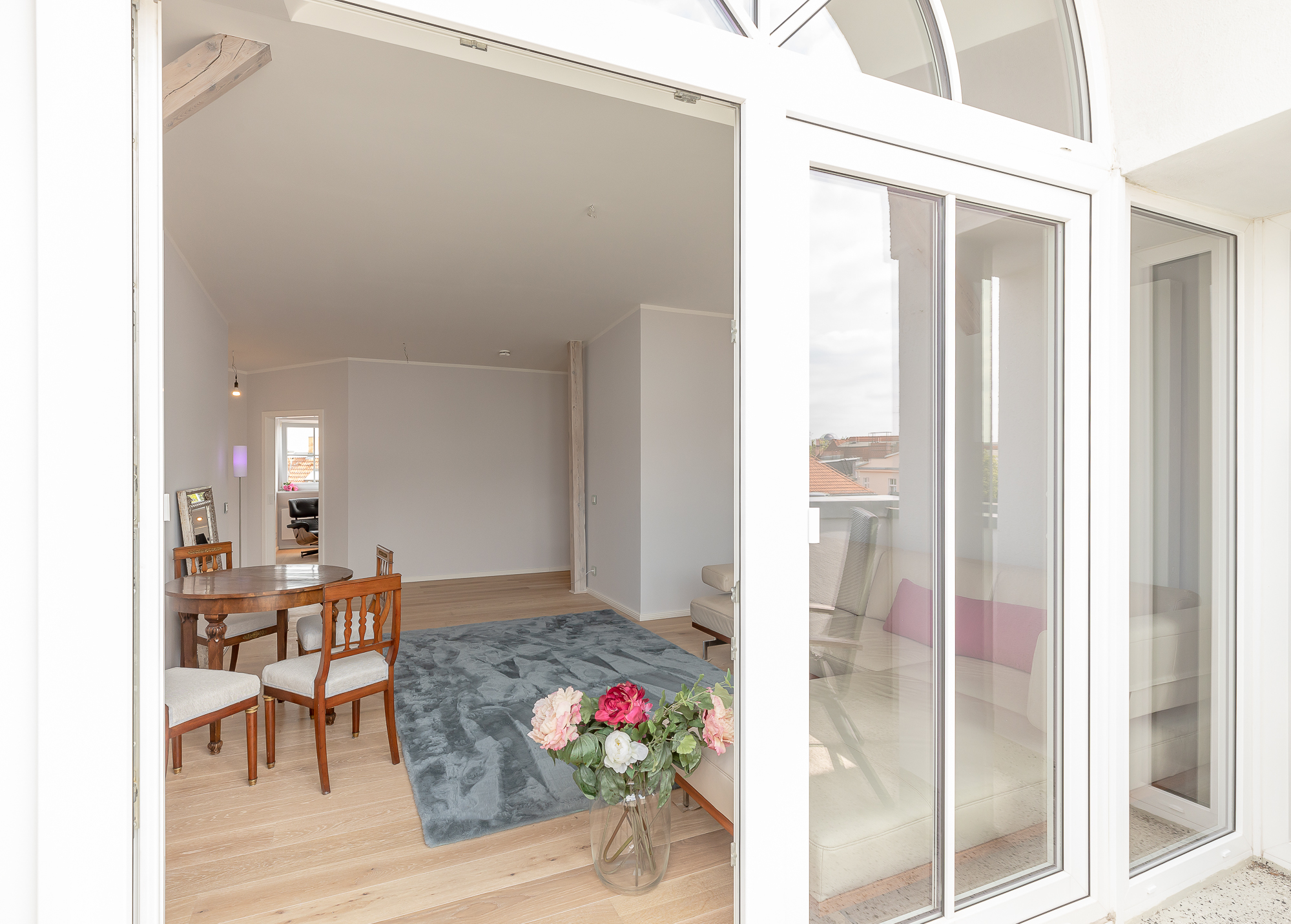 Helles Wohnzimmer mit Holzboden, Teppich, Esstisch, Stühlen und Glasvase mit Blumen.