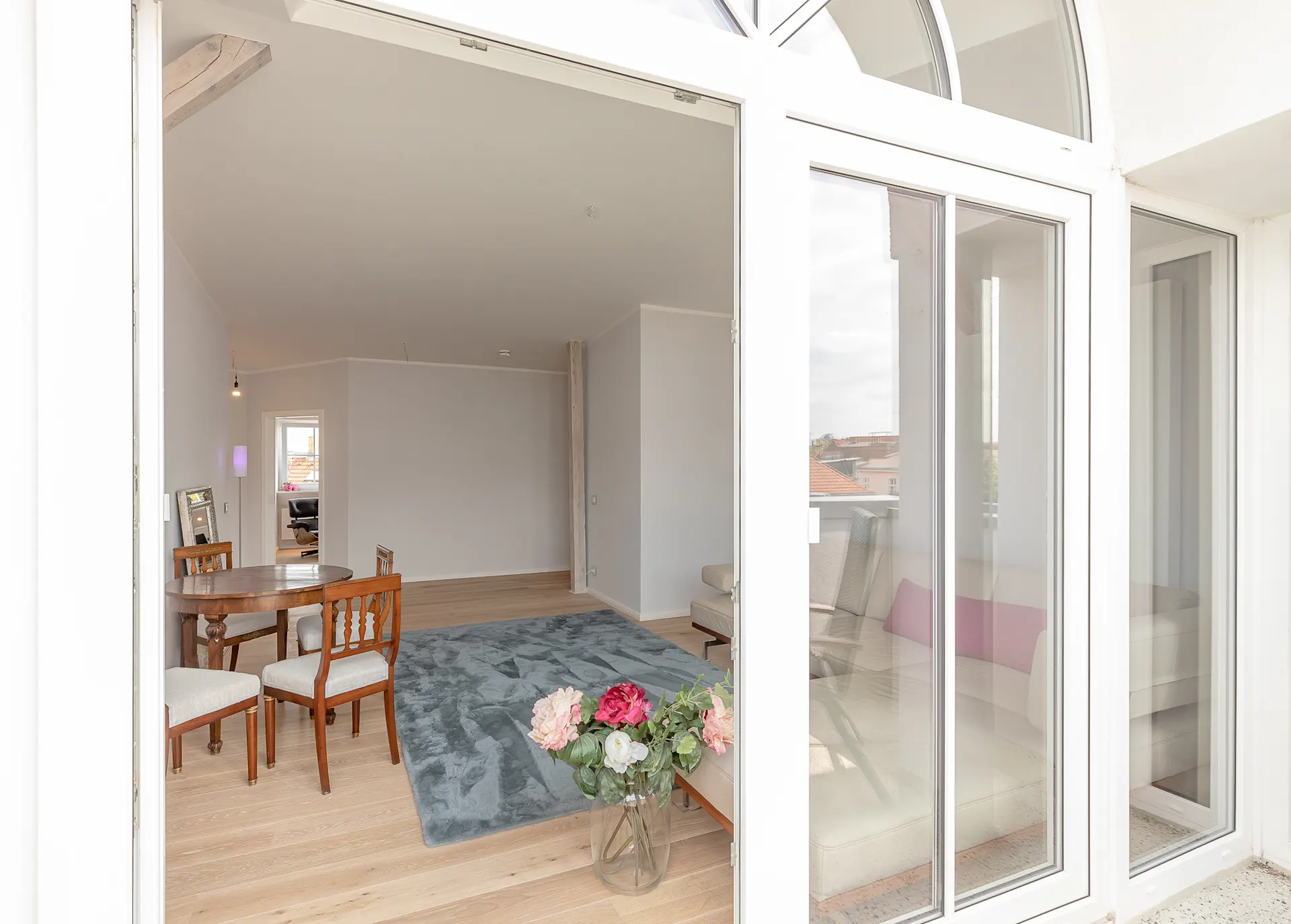 Helles Wohnzimmer mit Holzboden, Teppich, Esstisch, Stühlen und Glasvase mit Blumen.