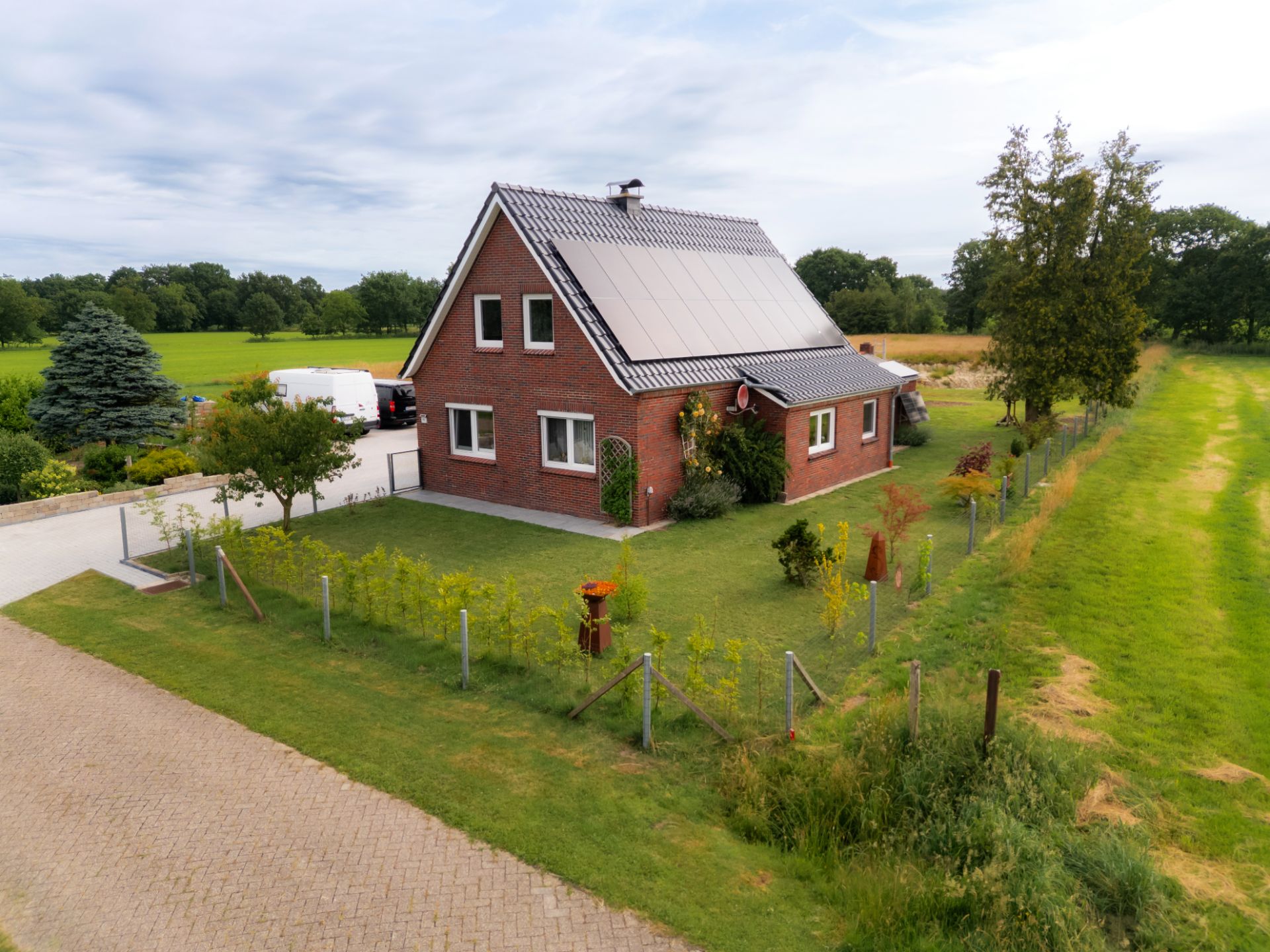 Backsteinhaus mit Solarpanels auf dem Dach und großem grünen Garten, umgeben von Feldern.