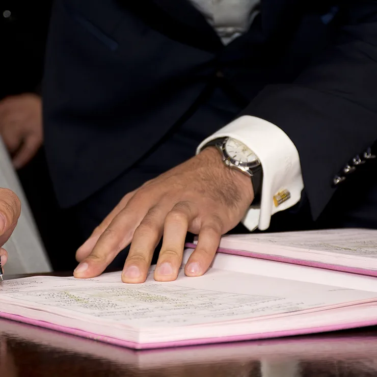 Nahaufnahme einer Person, die mit einem Stift ein Dokument auf einem Schreibtisch unterschreibt.